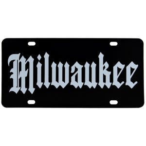 May include: Black license plate with the word "Milwaukee" in a gothic font. The text is light gray and centered on the plate. The plate has four mounting holes, one in each corner.