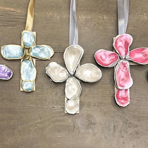 May include: Five decorative crosses with ribbon hangers. The crosses feature a variety of colors, including purple, blue, white, and pink. Each cross has a metallic gold or silver trim. The crosses are displayed on a wooden surface.