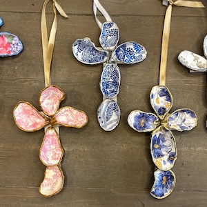 May include: Five decorative cross ornaments with floral and patterned designs. Each cross is made of fabric and has a ribbon for hanging. The crosses feature various colors, including pink, blue, and white, with gold trim.