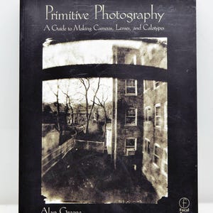 May include: A monochrome book cover for "Primitive Photography: A Guide to Making Cameras, Lenses, and Calotypes." The image shows a sepia-toned view of buildings and trees. The author's name, Alan Greene, is at the bottom. Published by Focal Press.