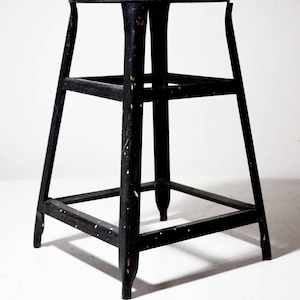 May include: A black wooden stool with a round seat and a square base. The stool has three supporting rungs and four legs. The black paint is distressed with white speckles, giving it a vintage look.