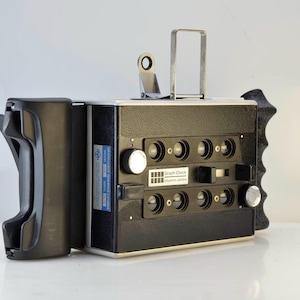 May include: A black and silver Graph-Check optics camera with multiple lenses and a handle. The camera has a rectangular shape with a silver metal handle on top and a black grip on the side. A blue and white label is on the side.