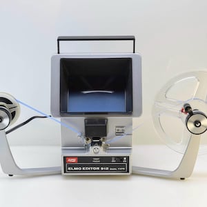 May include: Silver and black Elmo Editor 912 dual type film editor. The editor has a screen, two film reels, and a handle. The text "ELMO EDITOR 912 DUAL TYPE" is printed on the front.
