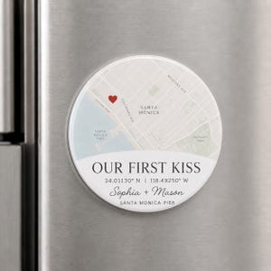 May include: A round magnet featuring a map of Santa Monica with a red heart icon. The text reads OUR FIRST KISS, 34.01130° N | 118.49250° W, Sophia + Mason, and SANTA MONICA PIER. The design includes street names like Ocean Ave and Colorado Ave.