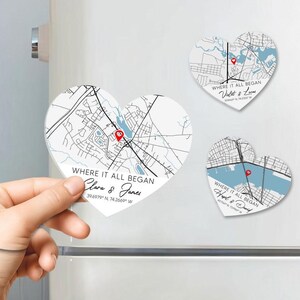 May include: Heart-shaped magnets featuring detailed map designs with a red pin marking a location. Each magnet has the phrase "WHERE IT ALL BEGAN" and names below. One magnet is held by a hand. The maps are in black and white.