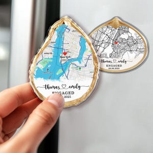 May include: Two oyster-shaped magnets with a gold trim. One magnet features a map of New York City with a red heart marking a location. The other magnet has a black and white map of New York City. Both magnets have the names "Thomas & Emily" and the word "ENGAGED" with the year 2025.