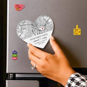 May include: A heart-shaped magnet with a map design and the text "OUR FIRST DATE richard & emily 40.70303°N, 73.97150°W 08.12.2026". The magnet is gray and white, with a red heart marking a location. Other magnets are also visible.