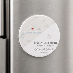 May include: A round white magnet featuring a map of Paris with a red heart icon, a gold ring icon, and the text ENGAGED HERE, 48.85660 N | 2.35220 E, Olivia & Noah, and 09.17.2022 printed in black and grey on a clean, minimalist background.