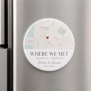 May include: A circular magnet featuring a map of Greenwich Village with a red heart icon. Below the map, the text reads WHERE WE MET, 40.74105 N | 73.98970 W, Emma & Lucas, and 05.12.2019. The design is printed on a white background.