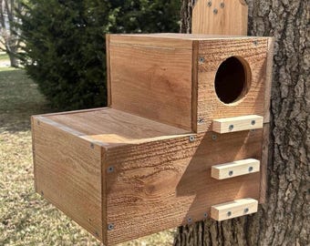 May include: A natural wood birdhouse with a stepped design, featuring a circular entrance hole on the upper level and three horizontal wooden slats attached to the front face. The structure is constructed with visible screws and mounted to a tree.