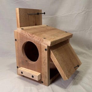 May include: A wooden birdhouse with a natural finish. The birdhouse has a circular entry hole, a slanted roof, and a hinged door. The birdhouse is constructed with screws and nails. The overall design is rustic and functional.
