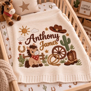 May include: A cream-coloured knit baby blanket featuring embroidered western motifs including a brown horse, a cowboy hat, a cactus, a wagon wheel, a lasso, and cowboy boots. The text "Anthony James" is embroidered in brown script in the centre.