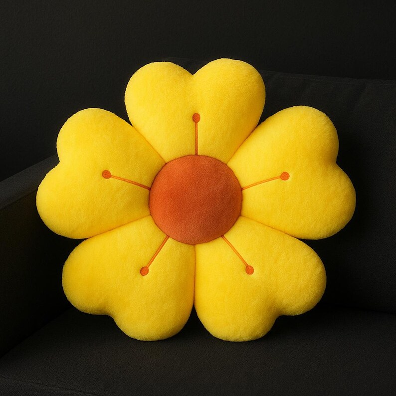 May include: A plush, yellow flower-shaped pillow with heart-shaped petals. The center is a contrasting orange-brown color. Orange stitching details radiate from the center to the tips of the petals. The pillow is on a black surface.