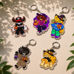 May include: Five acrylic keychains featuring cartoon characters in Halloween costumes. One wears a black hoodie with a pumpkin, another a pink shirt with text, a third a blue hoodie with a witch hat, a fourth a tan coat with a lantern, and a fifth a cape.