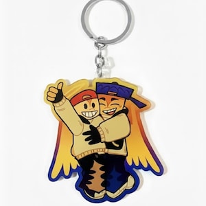 May include: A keychain featuring a cartoon illustration of two smiling figures embracing. One figure wears a red cap and the other a blue cap. They are dressed in tan sweaters and dark blue pants. The keychain has a silver ring and chain.