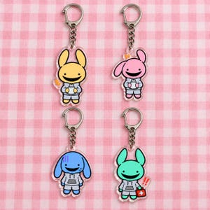 May include: Four acrylic keychains featuring cartoon rabbit-like characters in gray hoodies. One is yellow holding a cup, one is pink, one is blue with a worried expression, and one is green holding a red medical kit with a white cross symbol.