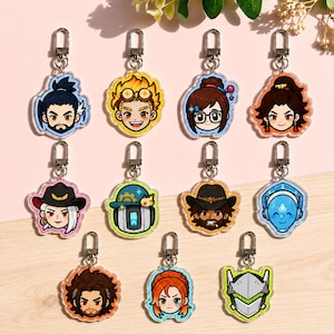 May include: A collection of eleven acrylic keychains featuring colorful chibi-style character portraits and helmets. Each charm includes a silver-toned metal lobster clasp attachment. The designs display various facial expressions, hairstyles, and headgear.