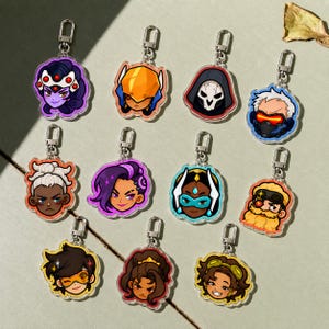 May include: A collection of eleven acrylic keychains featuring stylized, colorful character head illustrations from the video game Overwatch, each equipped with a silver-toned metal lobster clasp for attachment to bags or keys.