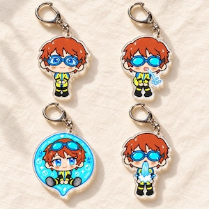 May include: Four acrylic keychains featuring a cartoon character with reddish-brown hair and blue goggles. The character wears a yellow and black wetsuit. Three keychains show the character in different poses, while one is circular with bubbles.