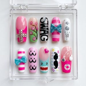 May include: A set of ten press-on nails in a clear plastic case featuring various designs including a rhinestone cross, cherry blossoms, the word SWAG, blue bows, a game controller, pink stripes, a mustache, a shark face, and a music player.