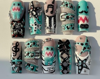 Op de afbeelding: Een verzameling van tien lange, versierde kunstnagels. Ontwerpen omvatten muzieknoten, sterren, strikken en cartoonfiguren, met accenten van zilveren kralen en glitter. Kleuren zijn teal, roze, zwart en wit.