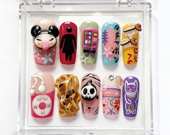 Könnte beinhalten: Ein Set aus zehn Acrylnägeln mit verschiedenen Designs, darunter eine Zeichentrickfigur, ein Verkaufsautomat, Sushi und eine Katze. Die Nägel werden in einer transparenten Kunststoffbox präsentiert.
