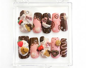 May include: A collection of ten artificial nails displayed in a clear case. The nails are decorated with chocolate, strawberries, and heart motifs. The colour scheme includes brown, pink, and white, with some nails having a textured, three-dimensional appearance.