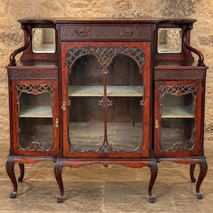 May include: A dark wood sideboard featuring a central glass-paneled cabinet flanked by two smaller glass-paneled cabinets. The piece includes a top drawer with ornate metal pulls, two small mirrors on the upper shelf, and curved cabriole legs.