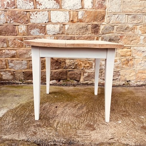 May include: A small wooden table with a light-coloured, rectangular top and white legs. The table is set against a brick wall. The table's dimensions are approximately 76 cm long, 51 cm wide, and 71 cm high.