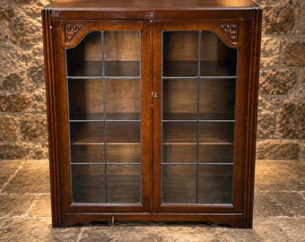May include: A dark brown wooden cabinet featuring two glass doors with a grid pattern. The cabinet has carved corner details at the top of each door frame and vertical fluted side panels. The interior contains three visible wooden shelves.