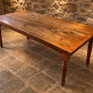 May include: A rectangular wooden dining table with a warm brown finish and visible wood grain. The table features a smooth, flat top surface and four sturdy legs with a turned, cylindrical design that tapers towards the bottom feet.