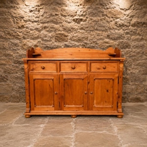 May include: A light brown wooden sideboard featuring three upper drawers with round knobs and three lower cabinet doors. The piece includes a decorative backboard with small side shelves and rests on short, rounded feet, showcasing a natural wood grain.