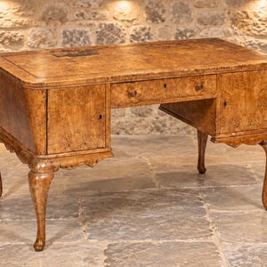 May include: A wooden desk featuring a light brown burl wood grain finish, a rectangular top with a dark inlay, a central drawer, and two side cabinets. The desk is supported by four curved cabriole legs with carved detailing at the top.