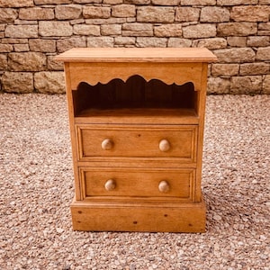 May include: A small, light brown wooden cabinet with two drawers and a shelf. The cabinet has a scalloped top edge and round wooden knobs on the drawers. It is set on a gravel surface against a brick wall.