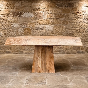 May include: A light brown wooden table featuring a thick, flat rectangular top with a natural wood grain pattern and a wide, tapered pedestal base. The table is constructed from solid wood with a smooth finish, showcasing the organic texture.