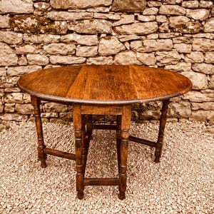 May include: An antique wooden drop-leaf table with a dark brown finish. The round table has turned legs and is set on a bed of small stones. The background features a stone wall. The table's diameter is approximately 90 cm.