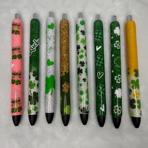 May include: A collection of eight pens with various designs. The pens feature glitter and patterns, including shamrocks, hearts, and the word "Lucky." Colors include pink, green, gold, and white. Each pen has a black tip.
