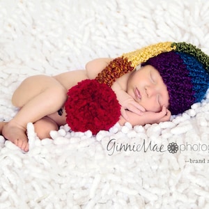 May include: A sleeping baby wearing a colorful crocheted hat with a red pom-pom. The hat has stripes of yellow, orange, red, blue, and purple. The baby is lying on a white fluffy blanket.