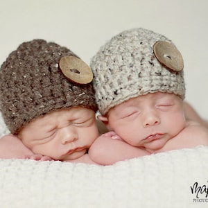 May include: Two newborn babies wearing brown and beige knitted hats with wooden buttons. The babies are sleeping on a white blanket.