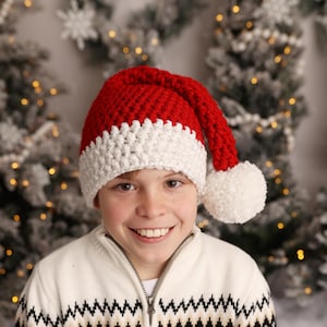 Op de afbeelding: Een jong persoon draagt een rood-witte gebreide kerstmanmuts met een witte pompon bovenop. De muts heeft een lange, puntige punt.