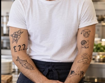May include: A plain white crew-neck t-shirt worn with a dark navy apron. The arms feature various black ink tattoos, including a flower, the number 22, a fish, a measuring cup with a globe, a snail, the text live fast, a hand holding a knife, and cherubs.