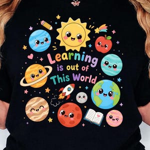 May include: A black t-shirt featuring a colourful graphic of smiling planets, a sun, a rocket, an apple, a pencil, and an open book. The text "Learning is out of This World" is printed in the centre in multi-coloured letters surrounded by small stars.