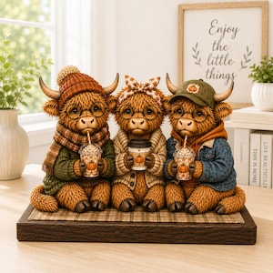 May include: Three brown highland cow figurines sitting on a wooden base. Each cow wears glasses and holds a cup with a pumpkin graphic. The left cow wears an orange knit hat and plaid scarf, the centre cow a patterned headband, and the right cow a green cap.