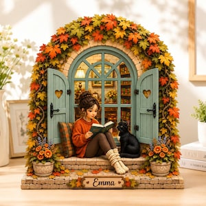 May include: A miniature diorama featuring a person in an orange sweater reading a book next to a black cat. The scene is framed by an arched window with light blue shutters, surrounded by autumn leaves. A plaque at the base displays the name Emma.