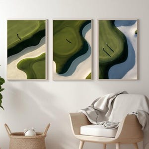 May include: Triptych of framed art prints showing aerial views of golf course greens and bunkers. The artwork uses a green and white palette, with golfers visible. The frames are light brown in colour.