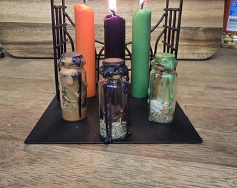 May include: A black metal candle holder with three lit candles in orange, purple, and green. Below the candles are three small glass bottles with dried herbs and wax drippings. The candles and bottles sit on a black metal base.