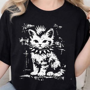 May include: A black t-shirt featuring a white graphic print of a kitten with a mohawk hairstyle and a spiked collar. The design has a distressed, textured appearance with rough edges, centered on the front of the garment with short sleeves.