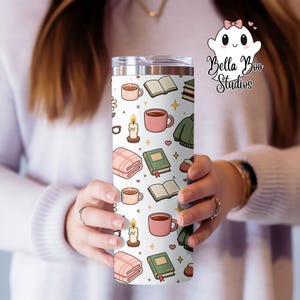 Peut inclure: Un gobelet blanc avec un couvercle en acier inoxydable, orné d'un motif d'illustrations chaleureuses. Le motif comprend des tasses roses, des livres, des bougies, des couvertures et un pull vert. Le gobelet est tenu par une personne.