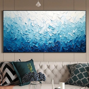 May include: Abstract oil painting in shades of blue and white. The artwork features textured brushstrokes, creating a sense of depth and movement. The painting is framed in a black border and hangs above a white sofa with decorative pillows.
