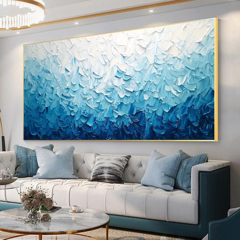 May include: Large abstract painting with a textured surface, featuring a gradient of blues and white. The artwork is framed in gold and hangs above a white sofa with blue pillows, creating a serene and modern living room aesthetic.